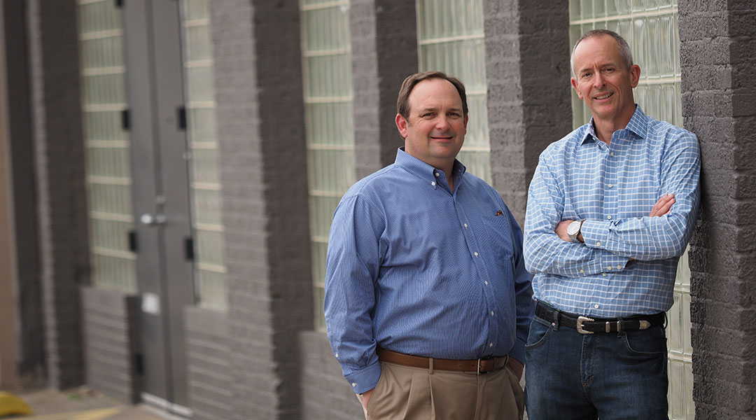 two men leaning on gray wall exterior shot blue button down shirts smiling business mensendero cross capital team geoff henrion and don at commercial real estate company in dallas texas